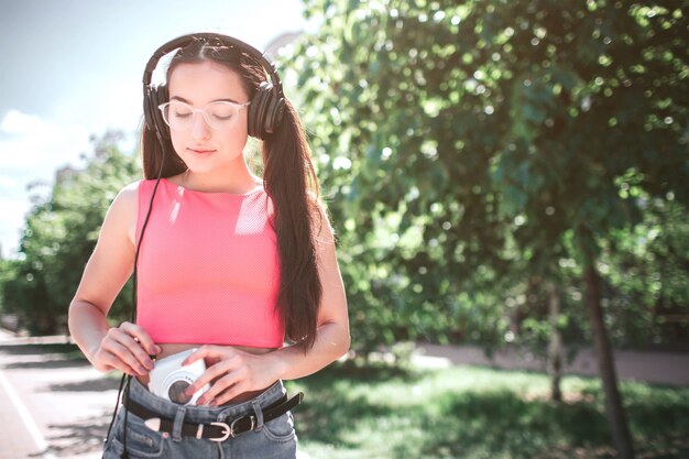 Una chica inteligente y hermosa a la que le gusta escuchar música está mirando al reproductor. Ella está conectando auriculares con reproductor blanco. Chica está parada en la calle.