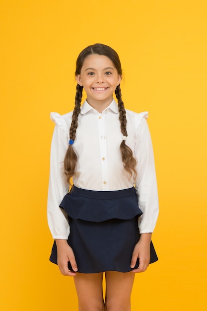 Foto chica inteligente linda niña sonriendo sobre fondo amarillo niña pequeña feliz vistiendo uniforme escolar niña de la escuela primaria con el pelo trenzado en largas trenzas adorable niña orgullosa de ser una alumna