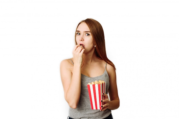 Una chica interesada viendo una película y comiendo palomitas de maíz en un blanco. La mujer mira con mucho cuidado la cámara.