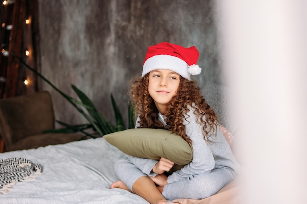 Chica interpolada hermosa de pelo rizado con sombrero de Santa y pijama sentado en la cama con la almohada, el tiempo de la mañana de Navidad