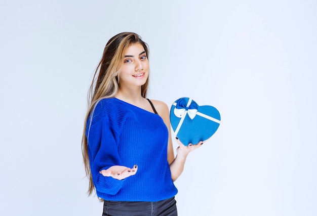 Foto chica invitando a alguien a presentar una caja de regalo con forma de corazón azul.
