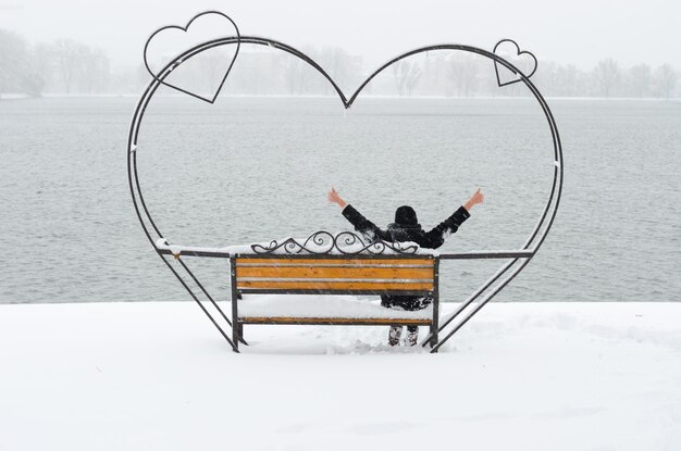 Chica irreconocible sentada en el hermoso banco de amor de hierro forjado en el parque con estanque y disfrutando de un hermoso día de invierno en soledad