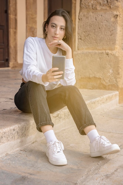 Chica italiana sentada en la calle con su teléfono celular mira fijamente a la cámara moda joven