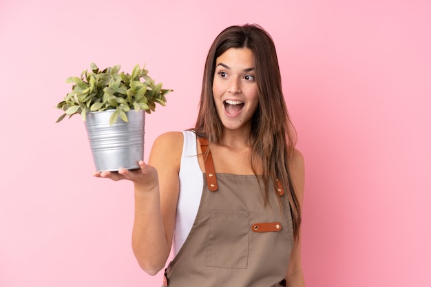 Chica jardinero adolescente sosteniendo una planta con sorpresa y expresión facial conmocionada