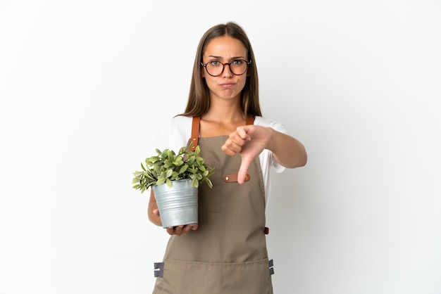 Chica jardinero sosteniendo una planta aislada mostrando el pulgar hacia abajo con expresión negativa