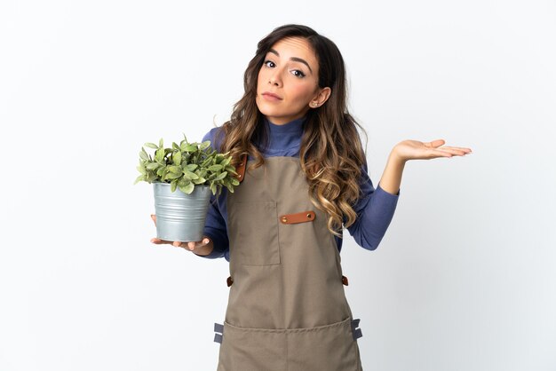 Chica jardinero sosteniendo una planta sobre fondo blanco con dudas mientras levanta las manos