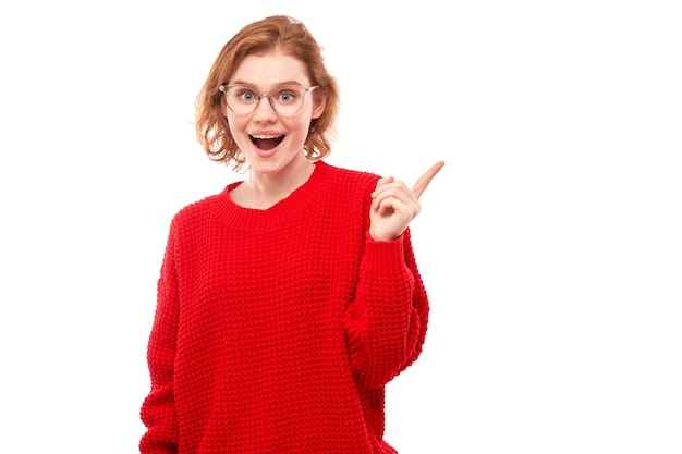 La chica de jengibre con cara de sorpresa en suéter rojo se ve sorprendida con la boca abierta en el fondo blanco del estudio señala con el dedo el espacio de la copia