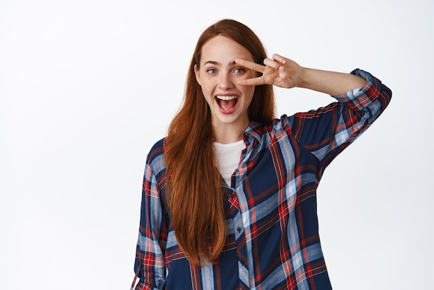 Chica de jengibre positiva riendo mostrando el signo de la paz cerca del ojo y sonriendo feliz de pie sin preocupaciones en camisa a cuadros contra el fondo blanco.