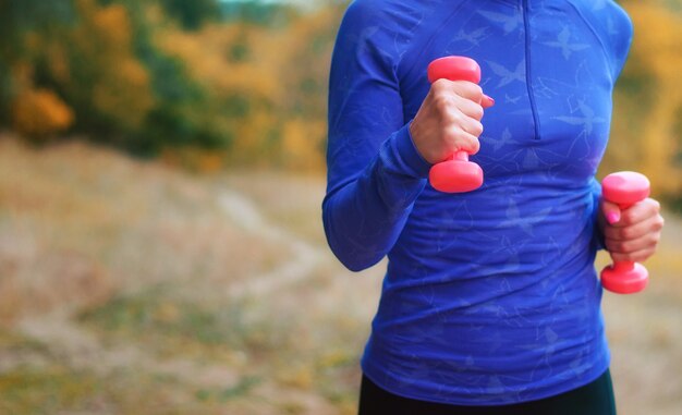 Chica jogger delgada en camisa azul con mancuernas rosas en sus manos realiza el ejercicio