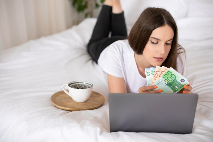 Chica joven acostada en una cama y sosteniendo moneda euro usa computadora portátil Trabajar desde casa