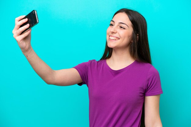 Chica joven aislada sobre fondo azul haciendo un selfie con teléfono móvil