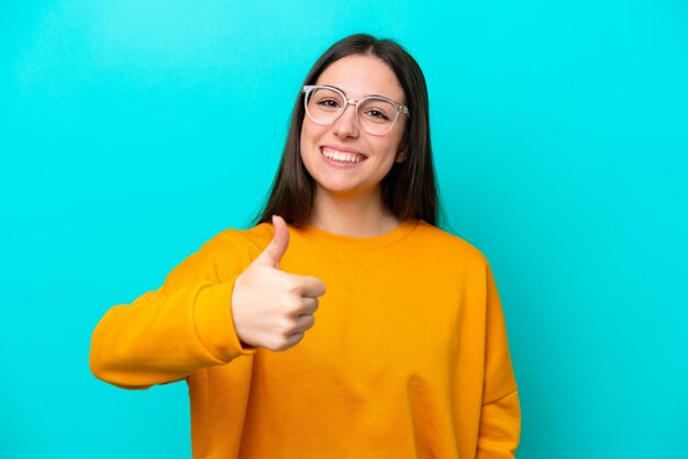 Chica joven aislado sobre fondo azul con gafas y con el pulgar hacia arriba