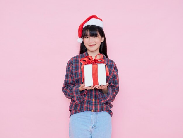 Chica joven alegre con gorro de Papá Noel con caja de regalo sobre fondo rosa