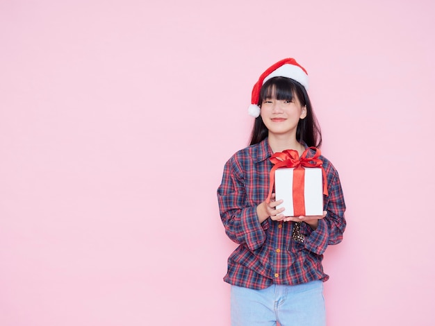 Chica joven alegre con gorro de Papá Noel con caja de regalo sobre fondo rosa