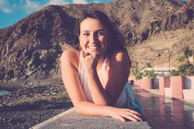 Chica joven alegre sonríe y disfruta del verano tendido al aire libre en actividades de ocio relajantes - montañas y playa en el fondo para la temporada de vacaciones