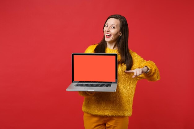 Chica joven alegre en suéter de piel amarilla apuntando con el dedo índice en la computadora portátil con pantalla vacía en blanco aislada sobre fondo rojo. Concepto de estilo de vida de emociones sinceras de personas. Simulacros de espacio de copia.