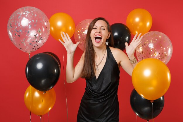 Chica joven alegre en vestidito negro gritando celebrando extendiendo las manos sobre globos de aire de fondo rojo brillante. Concepto de fiesta de día de San Valentín, día de la mujer, feliz año nuevo cumpleaños maqueta.