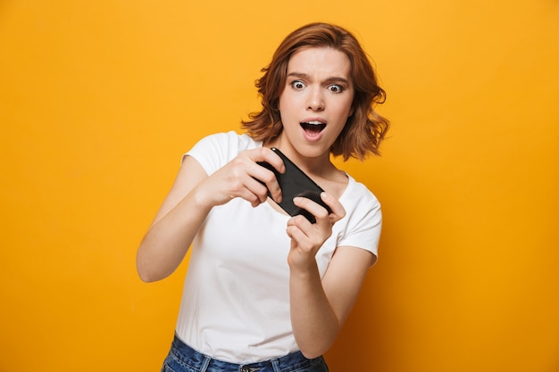 Chica joven alegre vistiendo camiseta que se encuentran aisladas sobre la pared amarilla, jugando juegos en el teléfono móvil