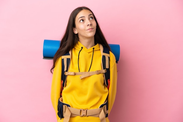 Chica joven alpinista con una mochila grande aislada sobre fondo rosa y mirando hacia arriba