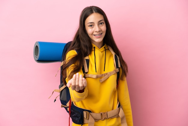 Chica joven alpinista con una mochila grande aislada sobre fondo rosa que invita a venir con la mano. Feliz de que hayas venido