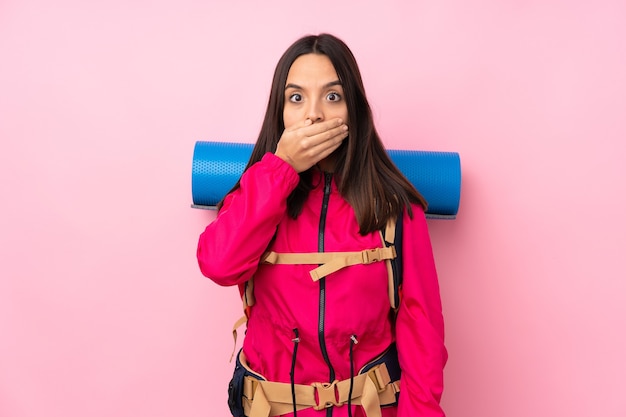 Chica joven alpinista con una mochila grande sobre rosa aislado cubriendo la boca con las manos