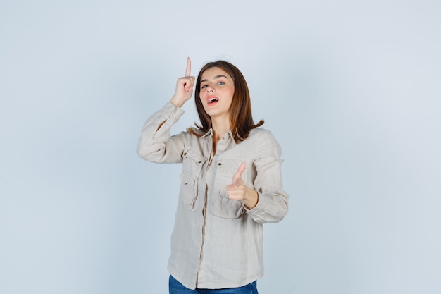 Chica joven apuntando hacia arriba con el dedo índice, mostrando el pulgar hacia arriba en camisa beige, jeans y luciendo linda vista frontal.