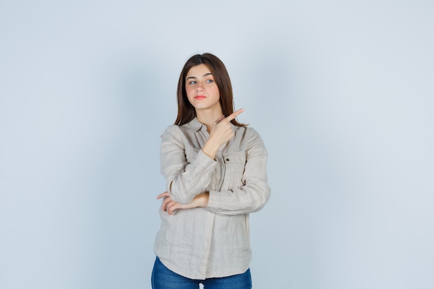 Chica joven apuntando a la derecha con el dedo índice en camisa beige, jeans y mirando alegre. vista frontal.