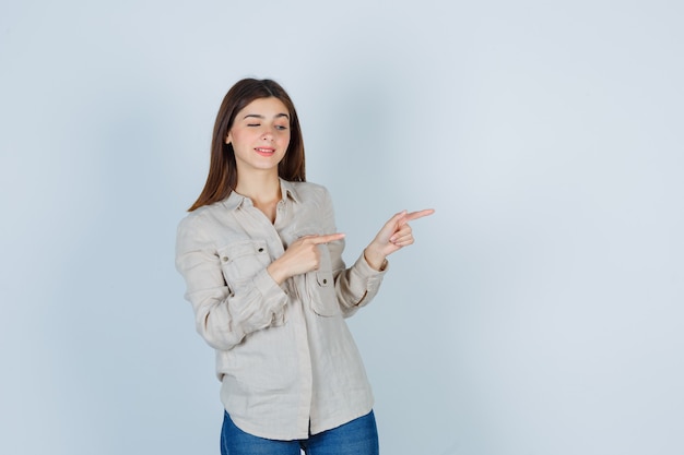 Chica joven apuntando a la derecha con los dedos índices en camisa beige, jeans y luciendo linda. vista frontal.