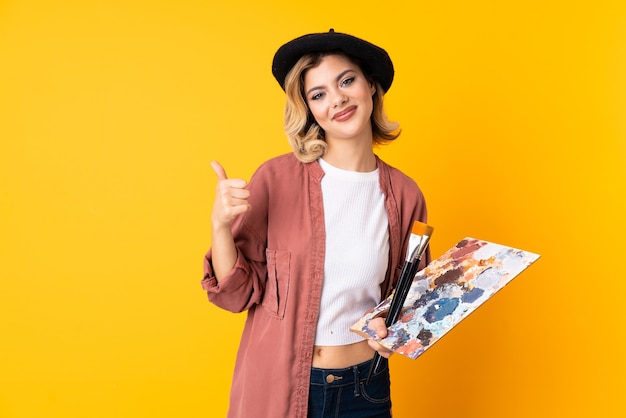 Chica joven artista sosteniendo una paleta aislada en amarillo dando un pulgar hacia arriba gesto