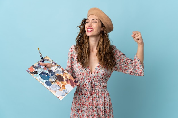 Foto chica joven artista sosteniendo una paleta aislada en la pared azul haciendo un gesto fuerte