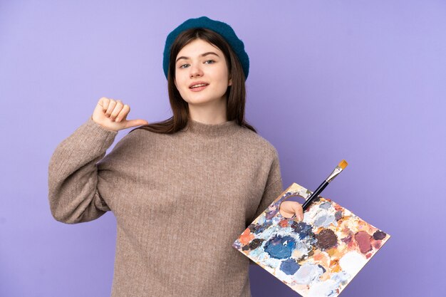 Chica joven artista sosteniendo una paleta sobre pared púrpura orgullosa y satisfecha