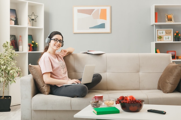 Chica joven de aspecto complacido con laptop usando audífonos y gafas sentado en el sofá detrás de la mesa de café en la sala de estar