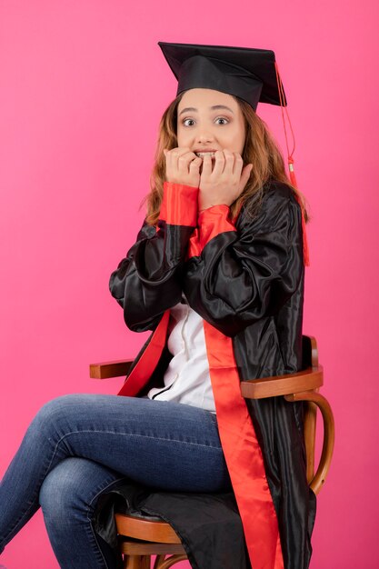 Chica joven asustada con toga de graduación y mordiéndose las uñas.