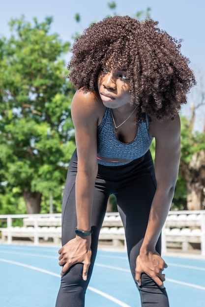 Chica joven atleta negra con cabello afro de pie con las manos apoyadas en las rodillas descansando después de una carrera mirando hacia el lado con pista de atletismo azul y árboles en el fondo