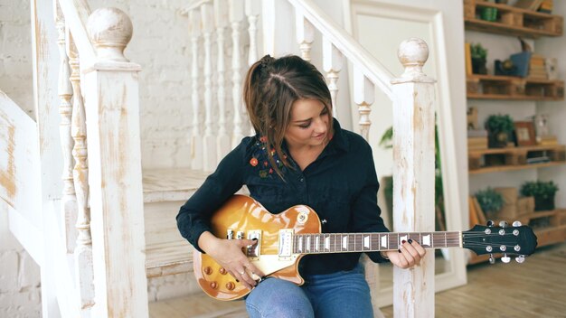 Chica joven atractiva aprendiendo a tocar la guitarra eléctrica sentada en las escaleras en el dormitorio en casa