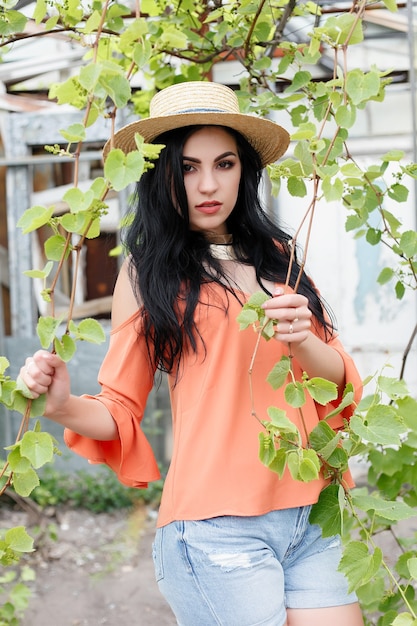 Chica joven atractiva con cabello ondulado con sombrero y traje elegante posando cerca de la vid