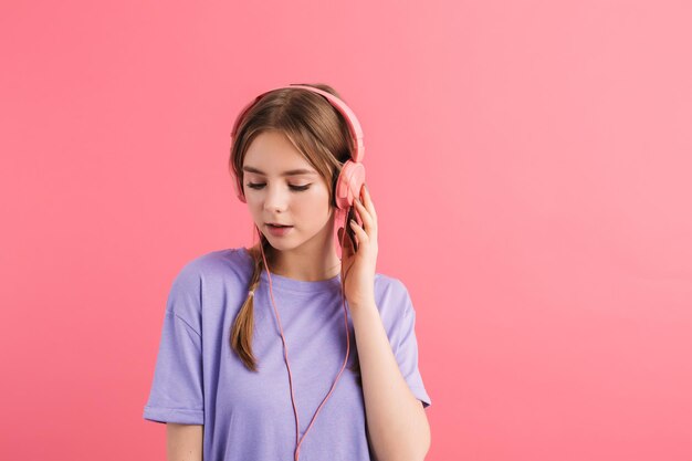 Chica joven atractiva con dos trenzas en camiseta lila escuchando música en auriculares mirando soñadoramente a un lado sobre fondo rosa aislado