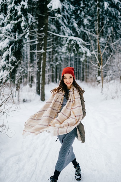 Chica joven atractiva hipster con una bufanda caliente caminando en el bosque de nieve
