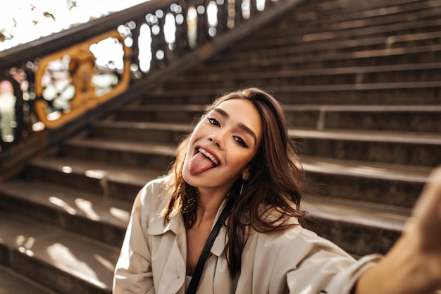 Chica joven atractiva con maquillaje de cabello ondulado moreno y gabardina beige sentada al aire libre haciendo selfie y haciendo muecas durante el cálido otoño soleado Fondo de escaleras de la ciudad vieja