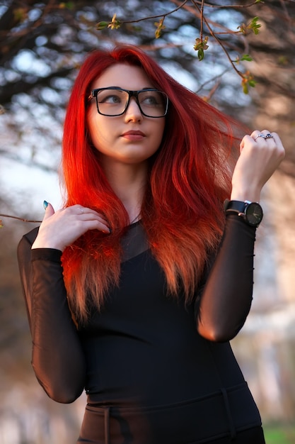 Chica joven atractiva con el pelo rojo, con gafas, posando cerca de un árbol