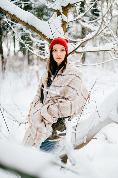 Chica joven atractiva que cubre con una capa caliente posando entre ramas de nieve