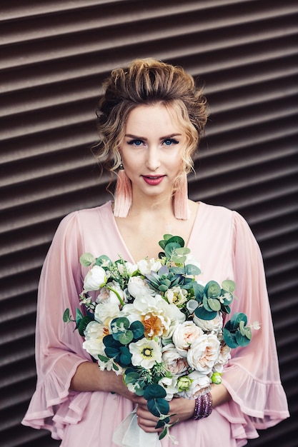 Chica joven atractiva en un vestido corto con un ramo de flores posando cerca de la pared de granito con persianas.