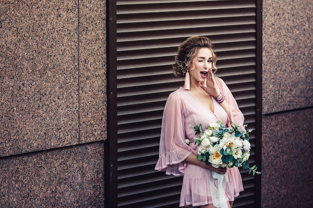 Chica joven atractiva con un vestido corto con un ramo de flores posando junto a la pared de granito con persianas enrollables.