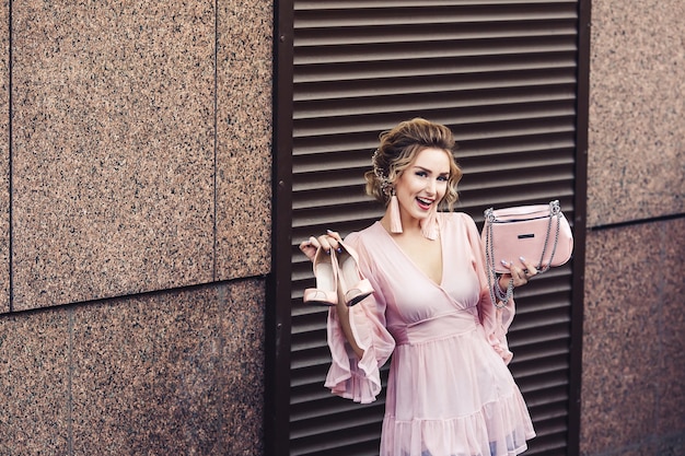 Chica joven atractiva con un vestido rosa corto sostiene los zapatos y la bolsa en la mano contra la superficie de las persianas enrollables. Concepto de compra y venta