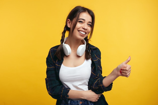 Chica joven con auriculares muestra como gesto aislado sobre pared amarilla