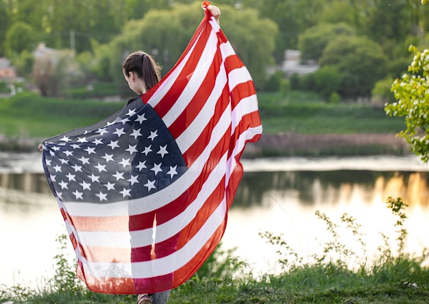 Chica joven con la bandera de América en la naturaleza desde atrás.