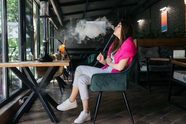 Chica joven bebiendo aperol Aperol spritz cóctel en un vaso La chica fuma una cachimba Cachimba en un restaurante Mujer fuma una cachimba y se sienta en una silla