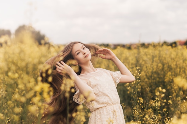 Chica joven y bella con el pelo largo volando en el viento con el telón de fondo del campo de colza. Brisa jugando con el cabello de la niña