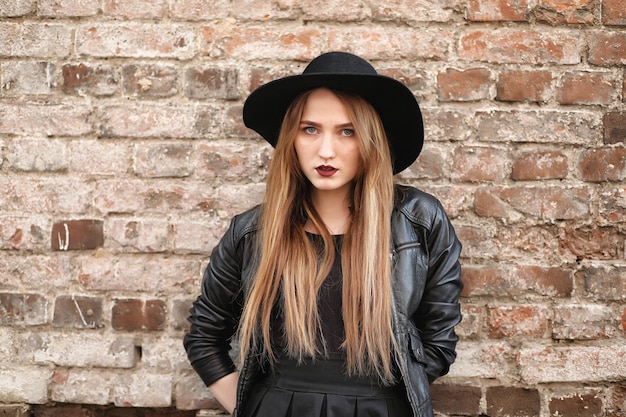 Chica joven y bella con un sombrero y con un exterior de maquillaje oscuro. Chica de estilo gótico en la calle. Una niña camina por las calles de la ciudad con un chaleco de cuero y un teléfono.
