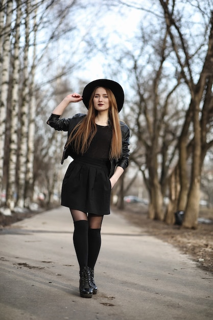 Chica joven y bella con un sombrero y con un exterior de maquillaje oscuro. Chica de estilo gótico en la calle. Una niña camina por las calles de la ciudad con un chaleco de cuero y un teléfono.